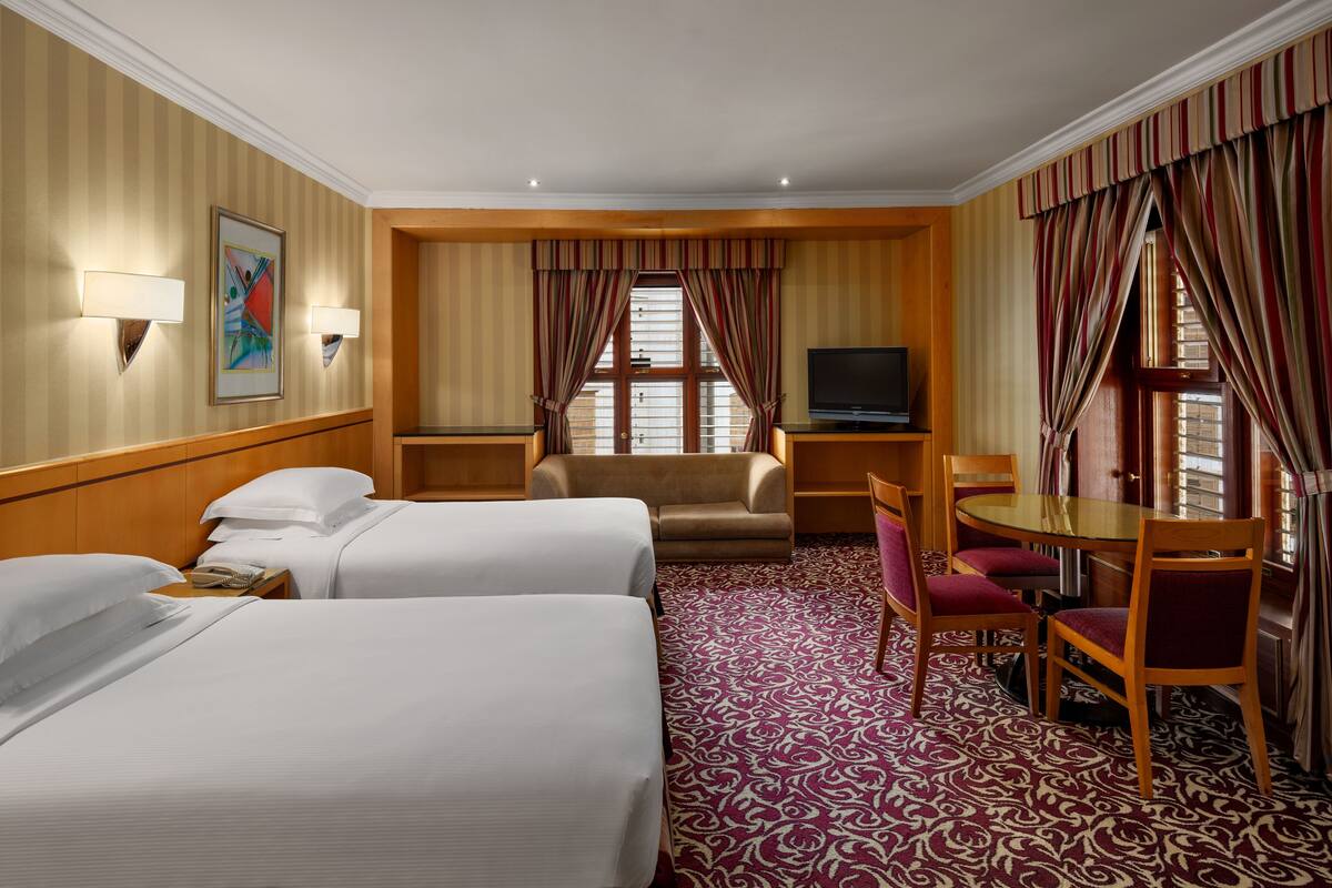 A hotel room with two beds, a TV, a dining table with chairs, and a window with curtains.