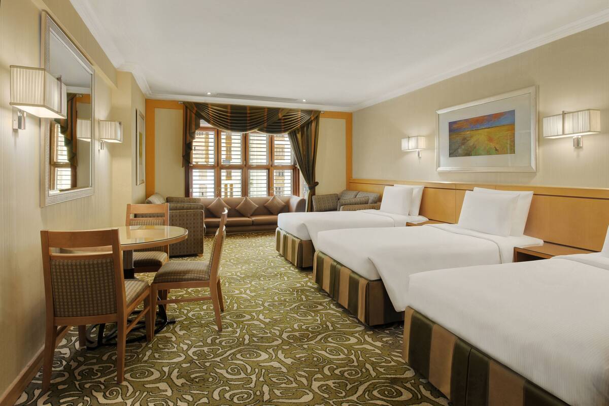 A hotel room with three beds, a dining table with chairs, a sofa, and a large window with curtains.