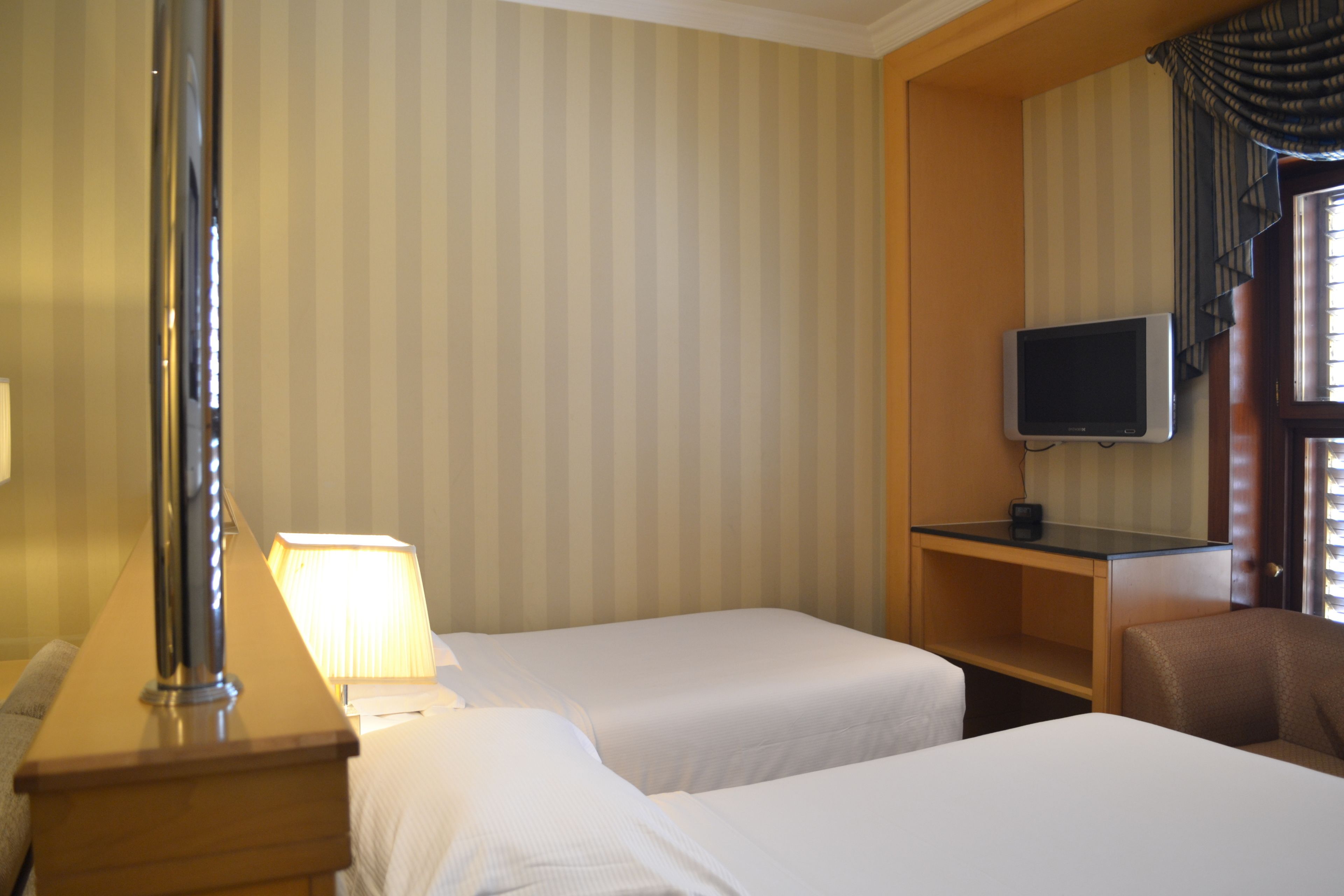 A hotel room with two beds, a desk with a lamp, a television on a stand, and a window with curtains.