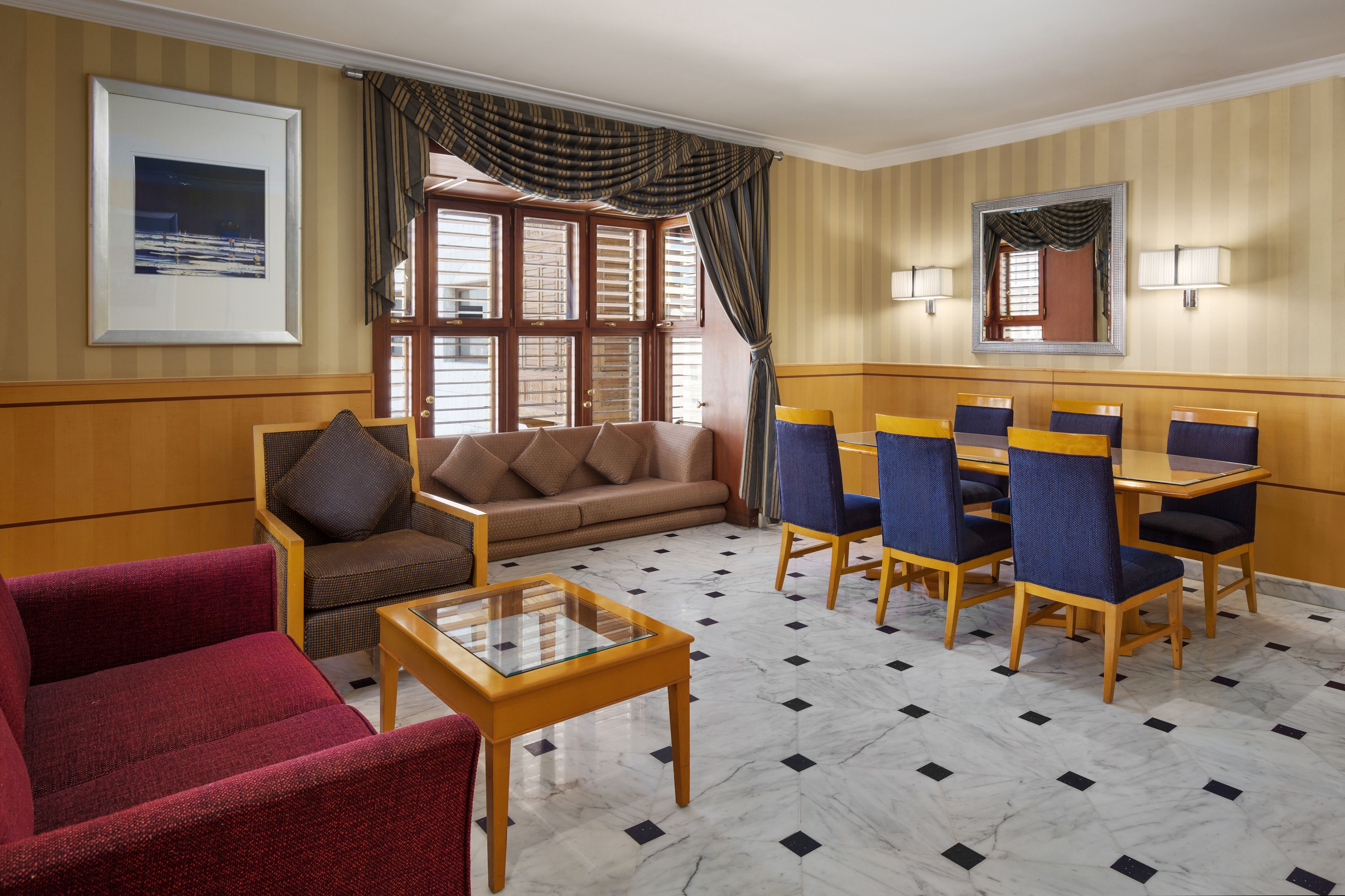 Habitación de hotel con sofá, sillón, mesa de comedor con sillas, una mesa de centro de cristal, un ventanal con cortinas y un cuadro en la pared.