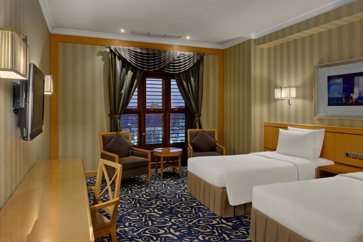 A hotel room with two beds, a desk, a chair, a small table, and a large window with curtains.