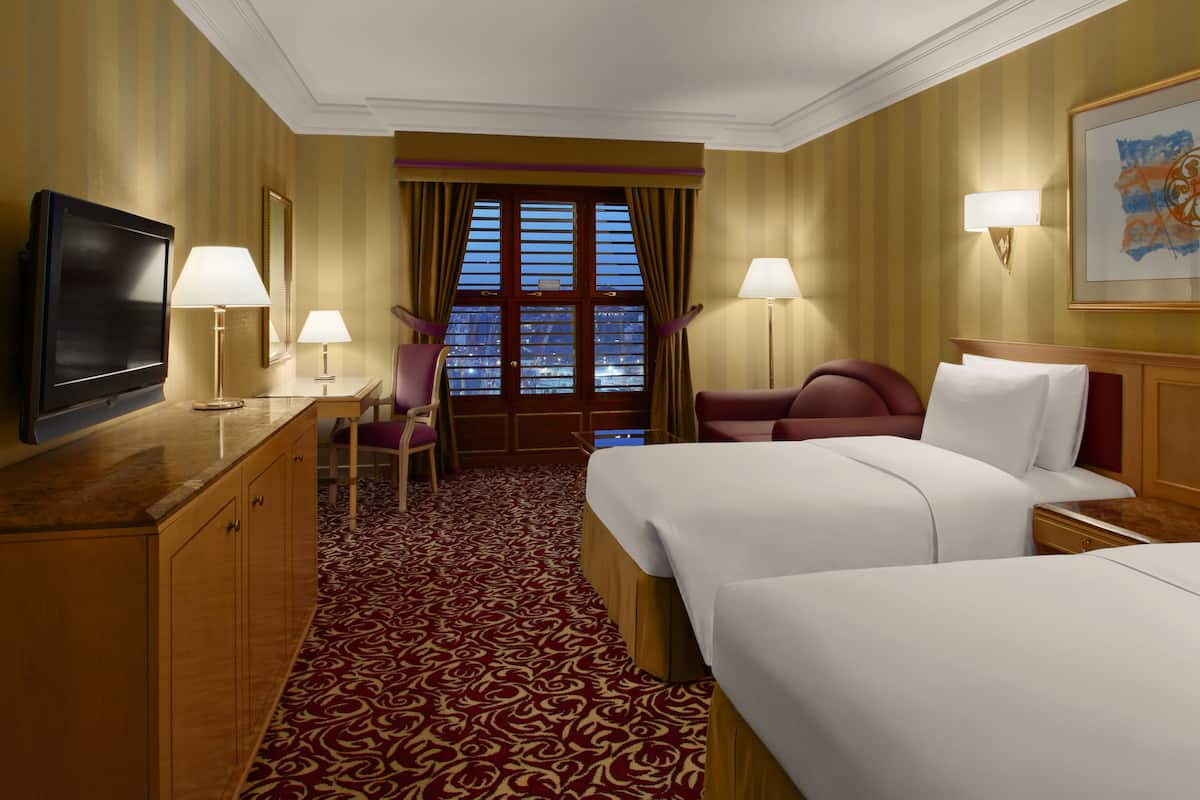 A hotel room with two beds, a TV, a desk with a lamp, a chair, and a window with curtains.