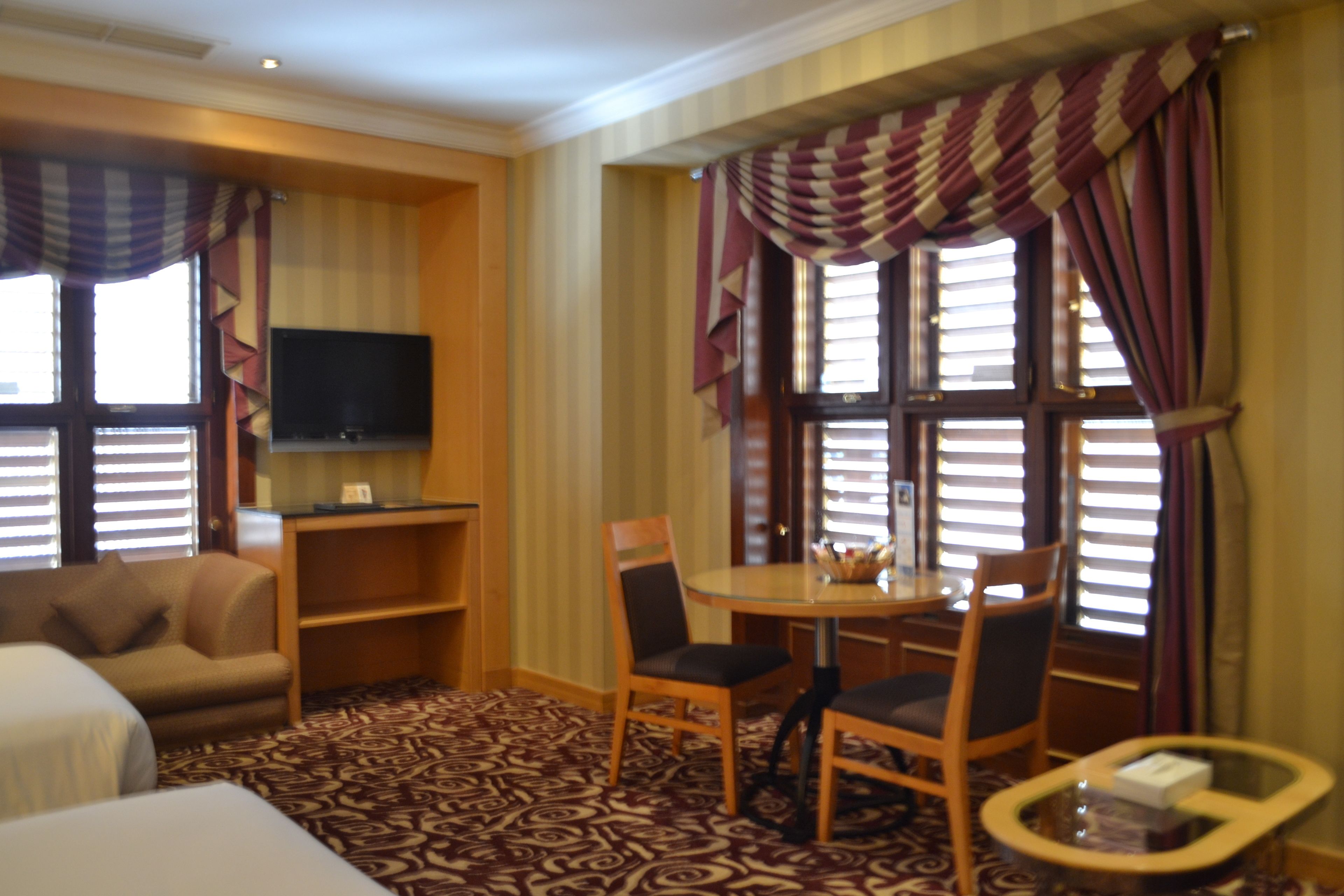 Habitación de hotel con sofá, una pequeña mesa con sillas, televisor montado en la pared y ventanas amplias con cortinas.