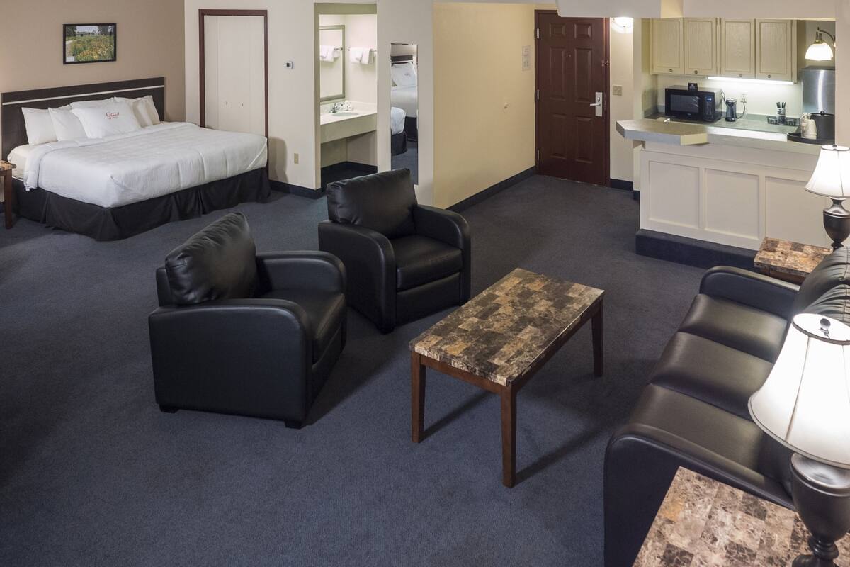 Una habitación de hotel con una cama, dos sillones de cuero negro, una mesita de madera, una lámpara y una kitchenette con microondas.