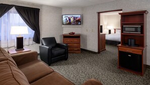 Ein Hotelzimmer mit einem braunen Ledersofa, einem schwarzen Sessel, einer Holzkommode, einem an der Wand montierten Fernseher und einer Mikrowelle auf einem Holzregal.