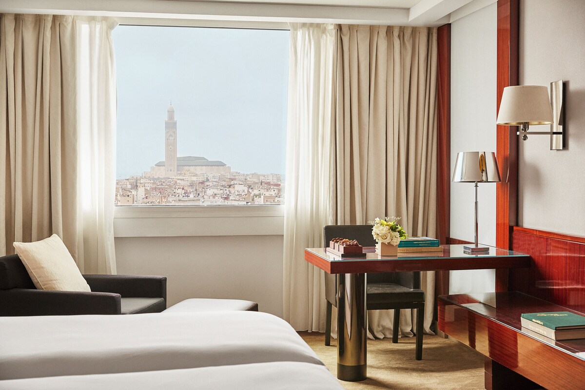 A hotel room with a bed, a desk with a lamp, a chair, and a view of a cityscape with a tower.