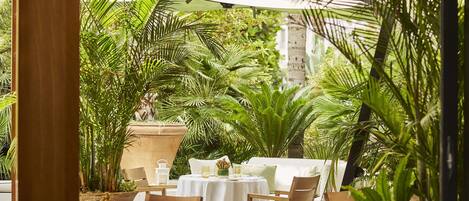 An outdoor dining area with tables and chairs, surrounded by lush greenery and a large umbrella providing shade.