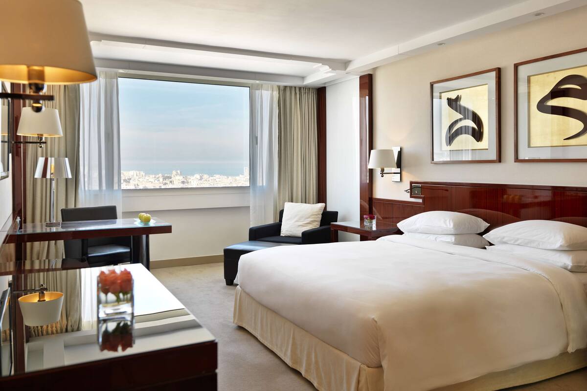 Une chambre d’hôtel avec un grand lit, un bureau avec une lampe, une chaise, une petite table sur laquelle est posé un vase de fleurs, et une vue sur la ville par la fenêtre.