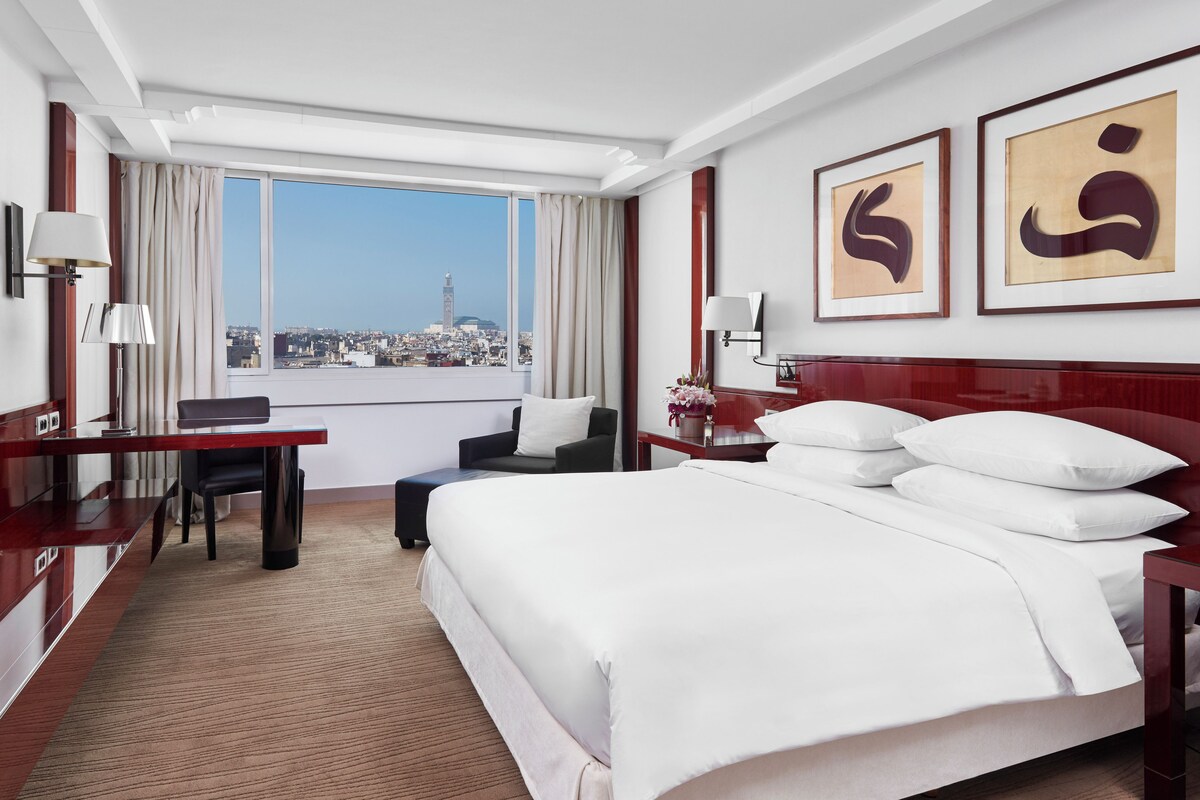 A hotel room with a large bed, a desk, a chair, and a view of a cityscape through a large window.