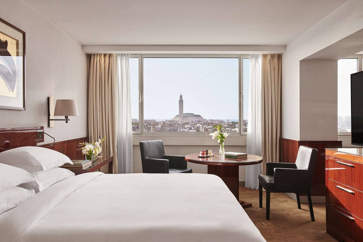 Une chambre d’hôtel avec un grand lit, deux fauteuils, une petite table ornée d’un vase de fleurs, un bureau avec une télévision et une vue sur le paysage urbain, avec une tour imposante en évidence.