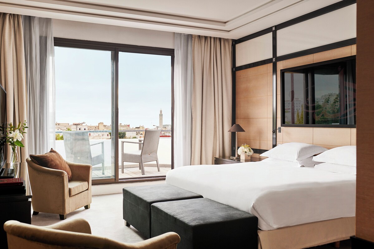 A hotel room with a large bed, a chair, a desk, and a view of the cityscape.