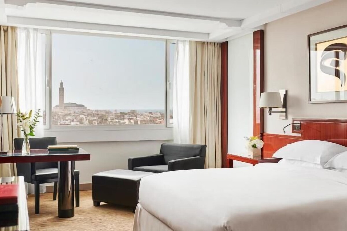 A hotel room with a bed, a desk, a chair, a nightstand, and a window with a city view.