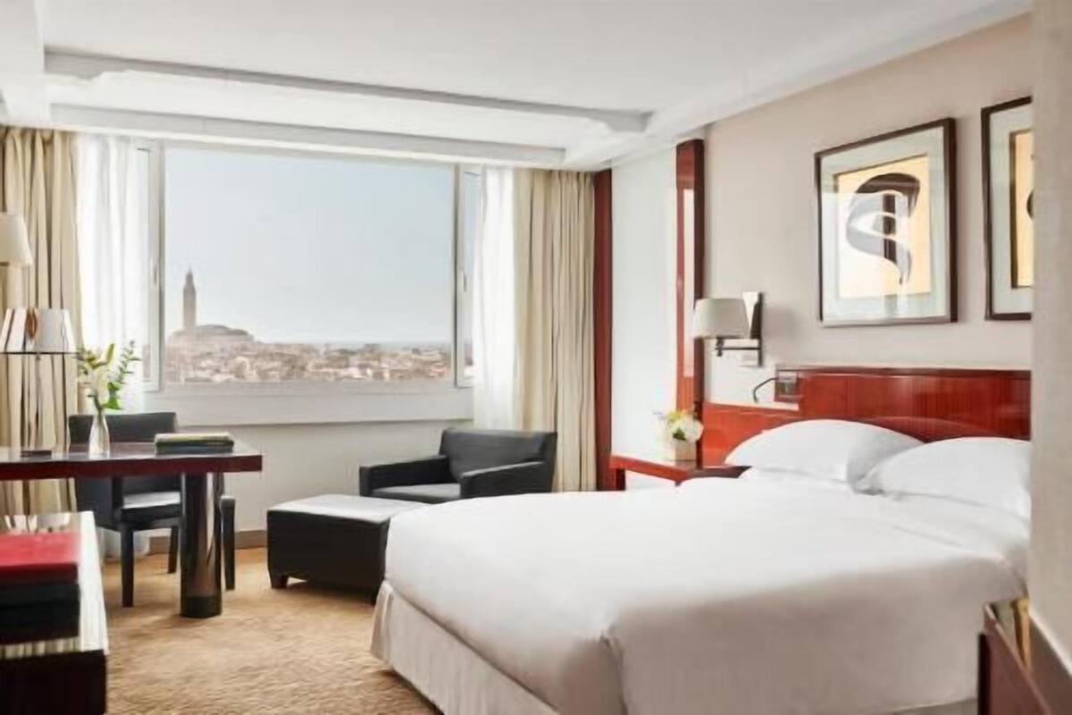 Une chambre d’hôtel équipée d’un lit, d’un bureau, d’une chaise, d’une table de chevet et d’une fenêtre donnant sur la ville.