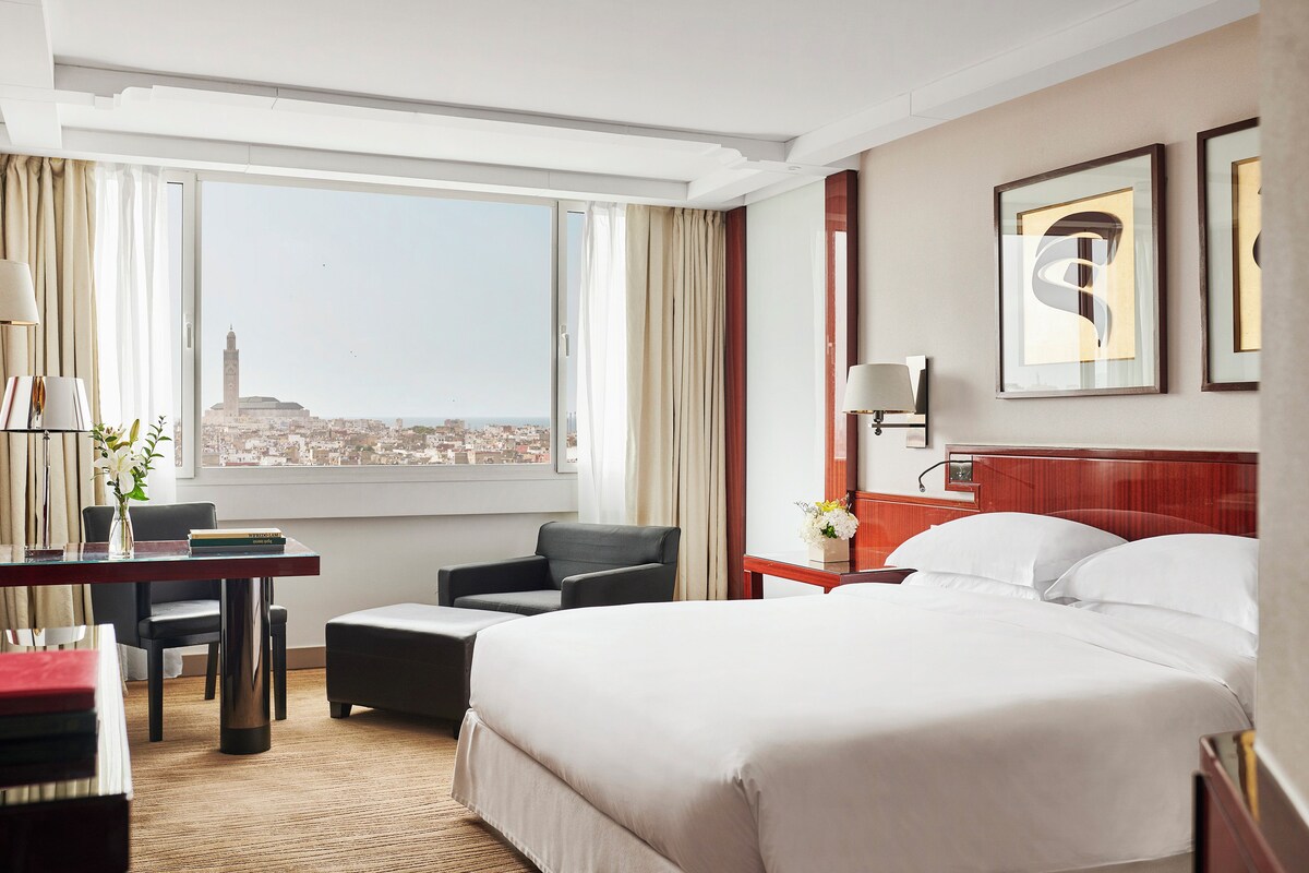 A hotel room with a large bed, a desk, a chair, and a view of a cityscape.