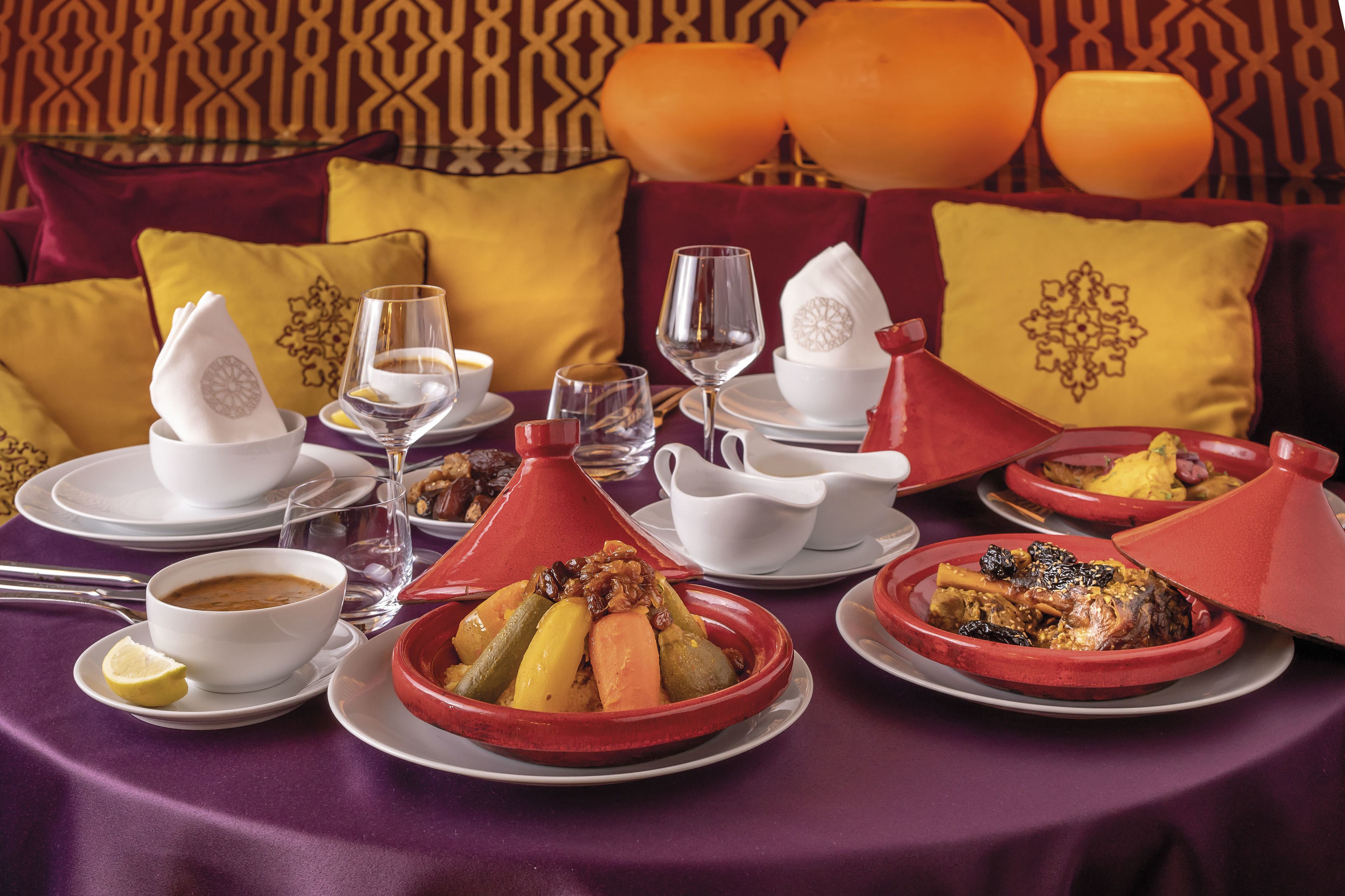 Una mesa de comedor dispuesta con cocina marroquí, incluyendo tajines y cuscús, en una habitación con papel tapiz estampado y cojines anaranjados.