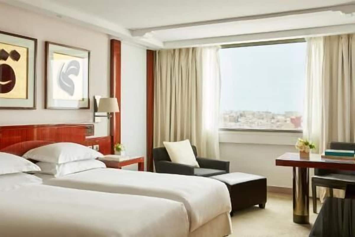 A hotel room with two beds, a desk, a chair, a lamp, and a view of a cityscape.