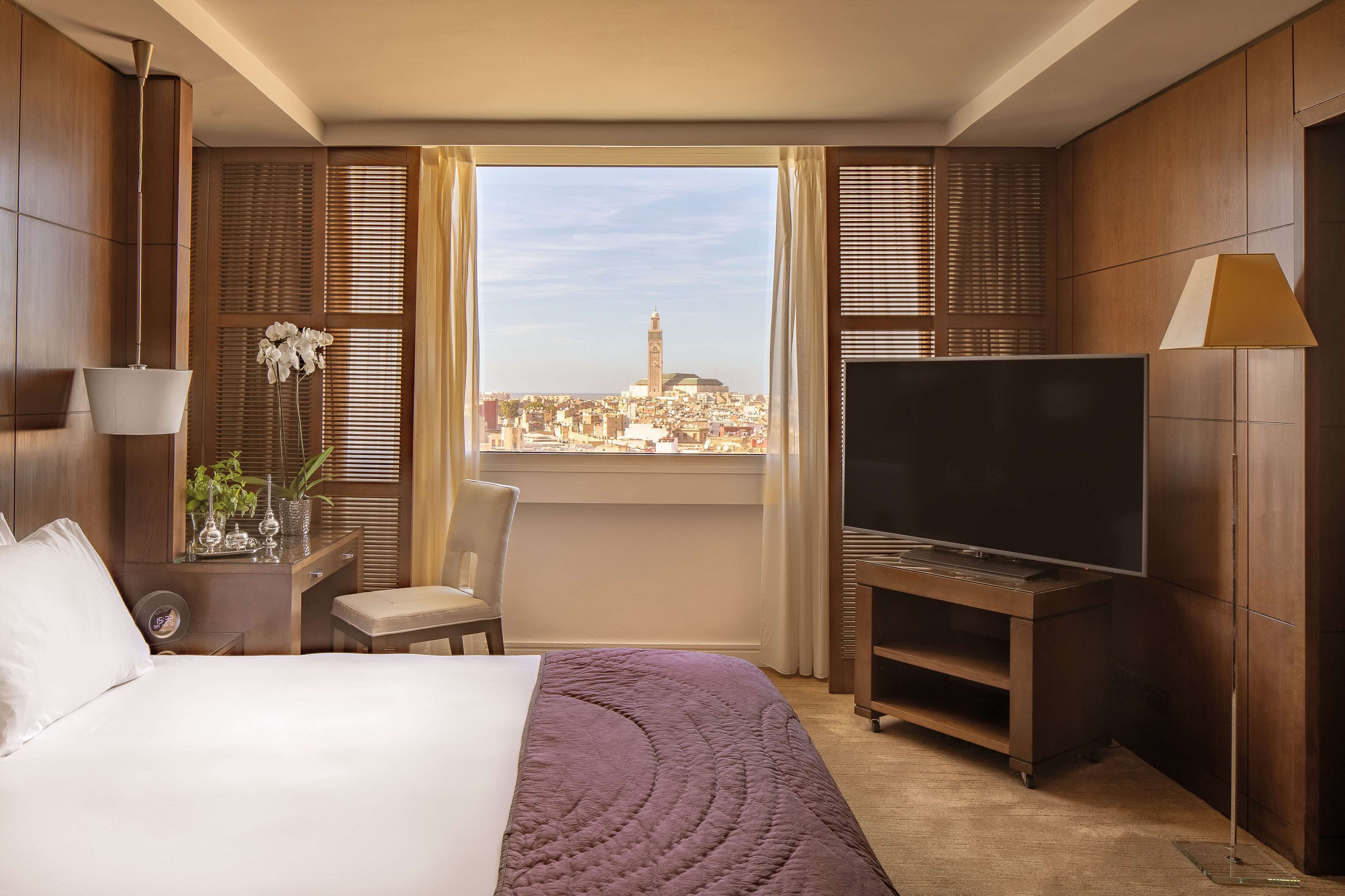 A hotel room with a bed, TV, and a view of a cityscape.