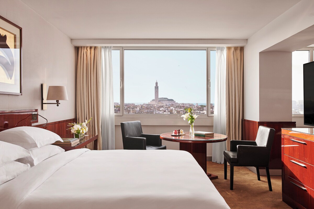 A hotel room with a bed, two chairs, a small table with a vase of flowers, a TV, and a view of a cityscape with a prominent tower.
