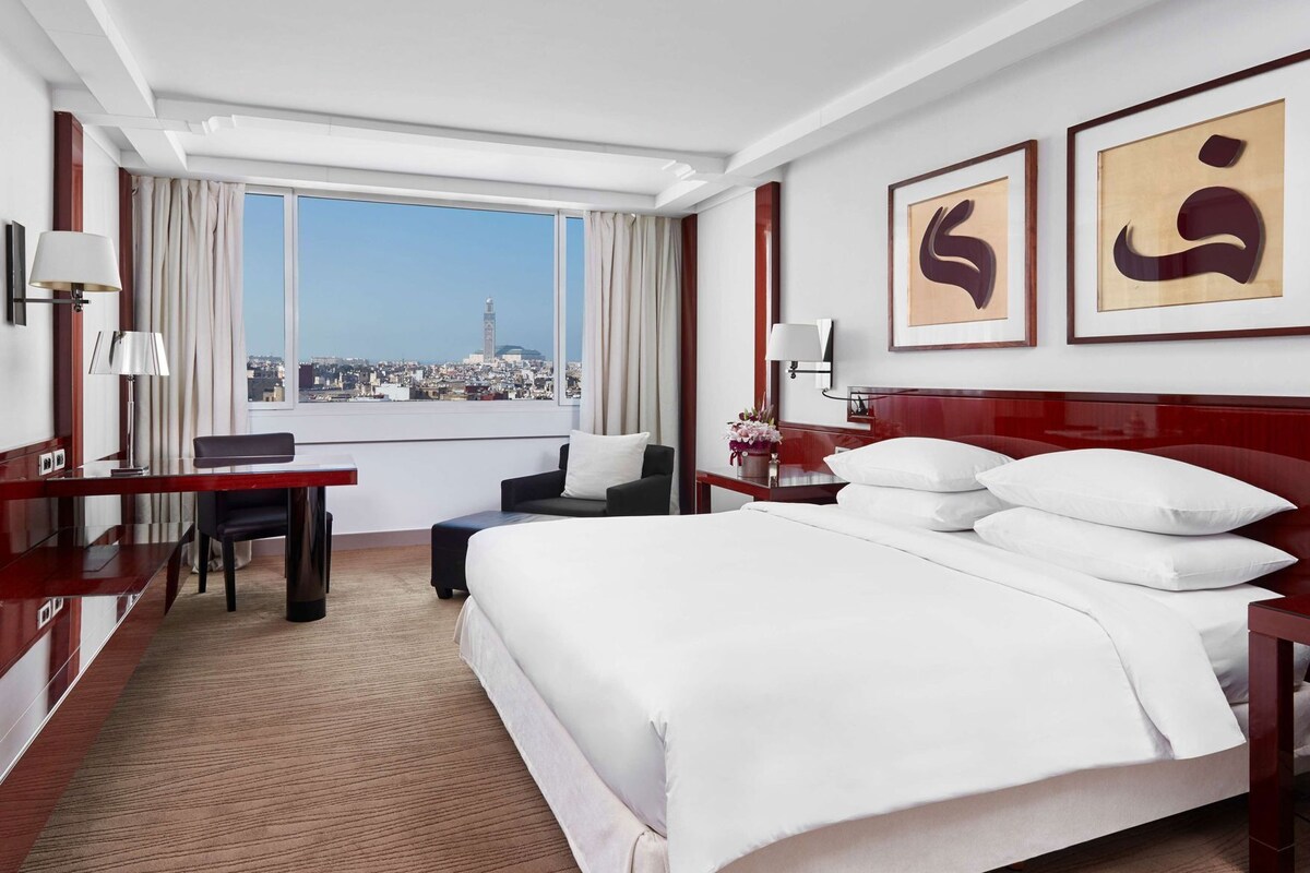 A hotel room with a large bed, a desk, a chair, a nightstand, and a view of a cityscape through a large window.