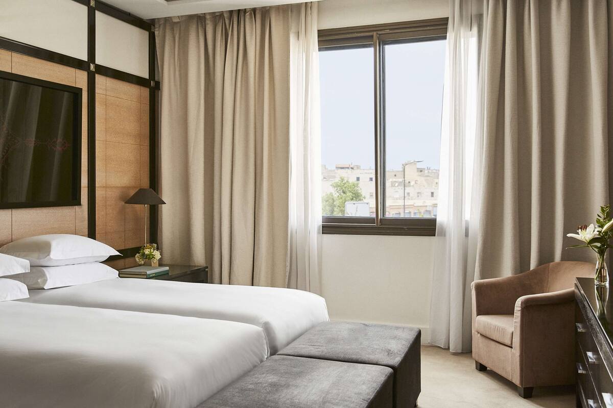 Une chambre d’hôtel avec deux lits, une chaise, une table de chevet avec une lampe, un vase de fleurs et une vue sur les bâtiments par la fenêtre.