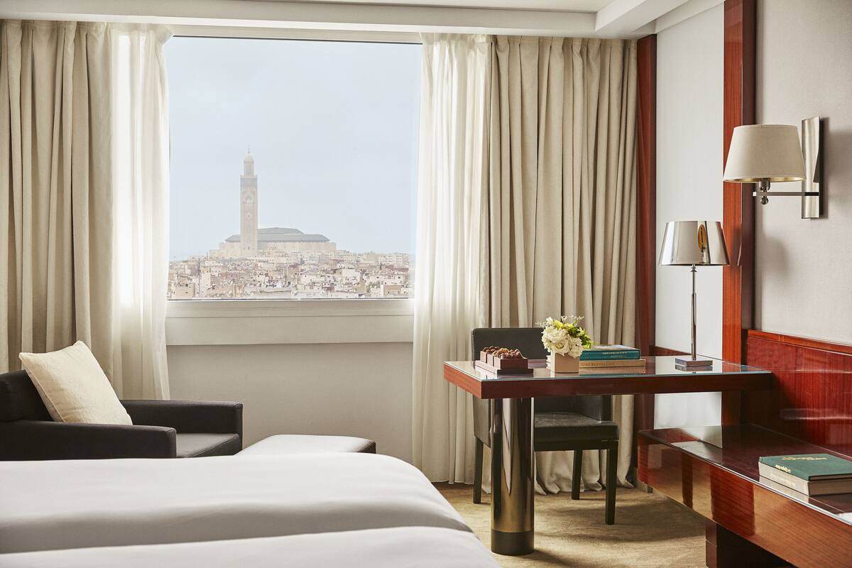 Une chambre d’hôtel avec un lit, un bureau avec une lampe, une chaise et une vue sur le paysage urbain, avec une tour en particulier.