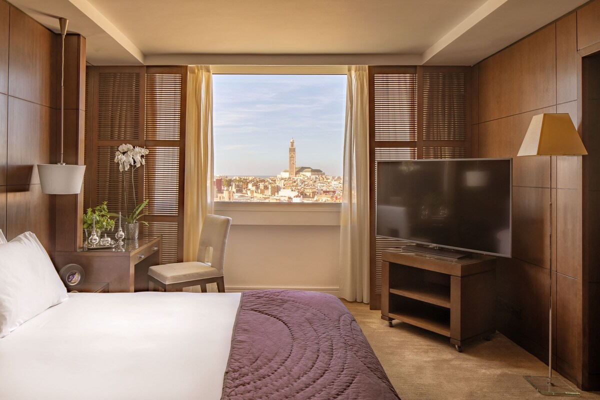 A hotel room with a bed, a chair, a TV, and a view of a cityscape.