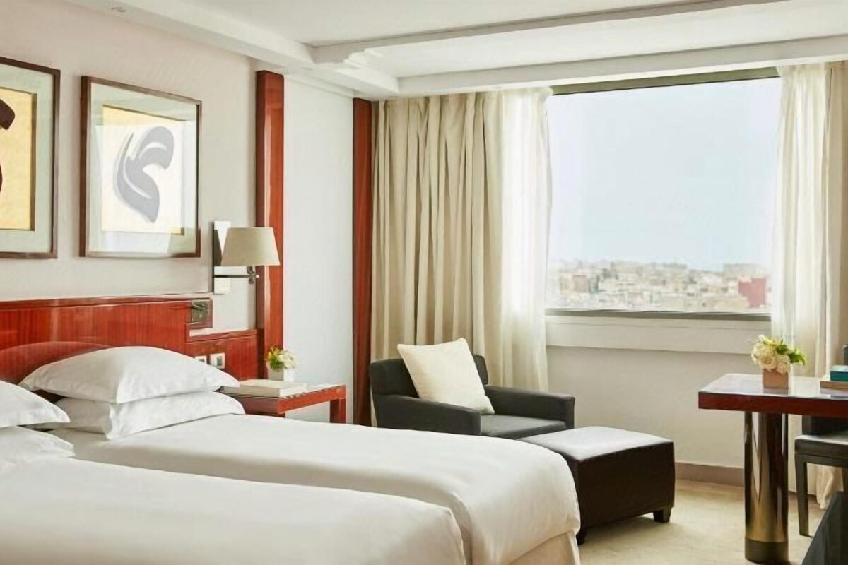 A hotel room with two beds, a desk, a chair, a view of the city, and artwork on the wall.
