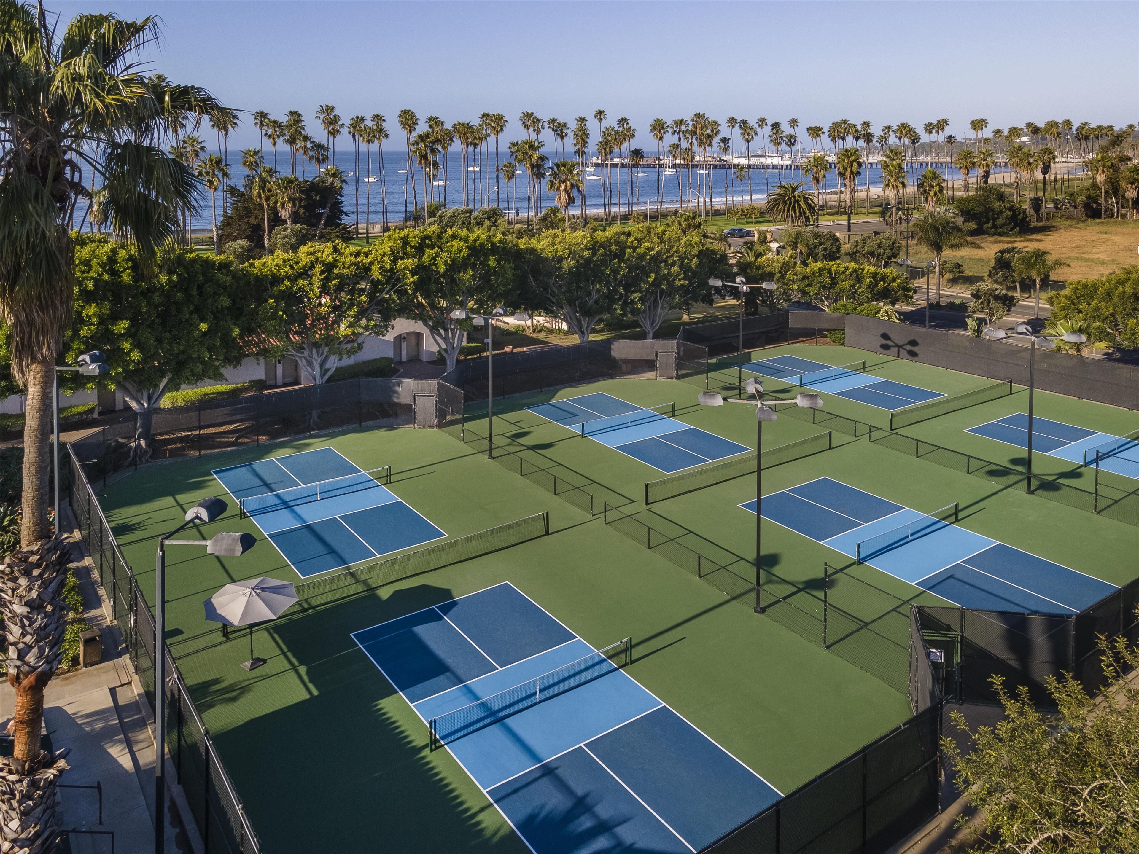 A tennis court complex with multiple courts, surrounded by palm trees and a view of the ocean.