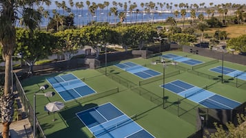 A tennis court complex with multiple courts, surrounded by palm trees and a view of the ocean.