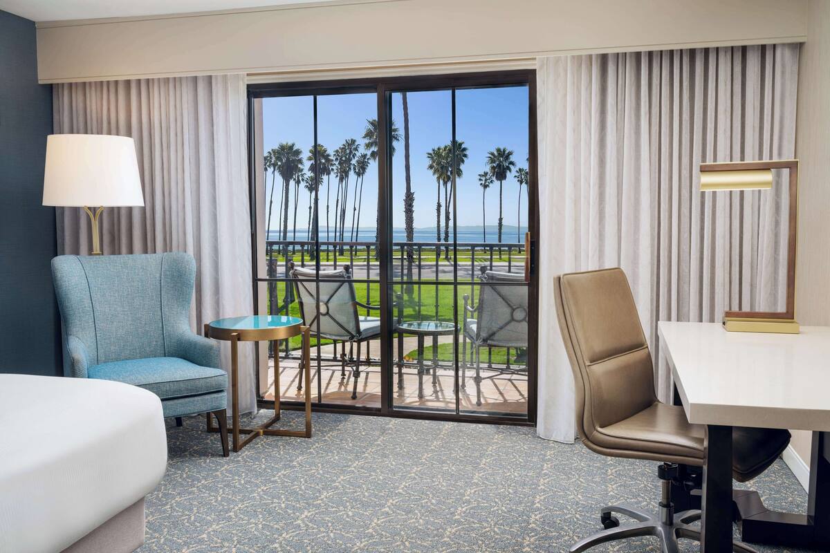 A hotel room with a bed, a blue armchair, a small table, a desk, and a view of palm trees and the ocean through a glass door.