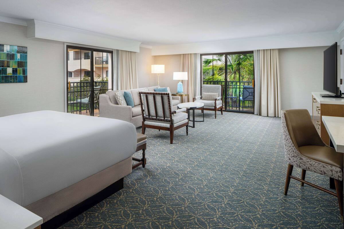 A hotel room with a large bed, a sofa, a chair, a desk, and a TV. There is a balcony with a view of greenery.