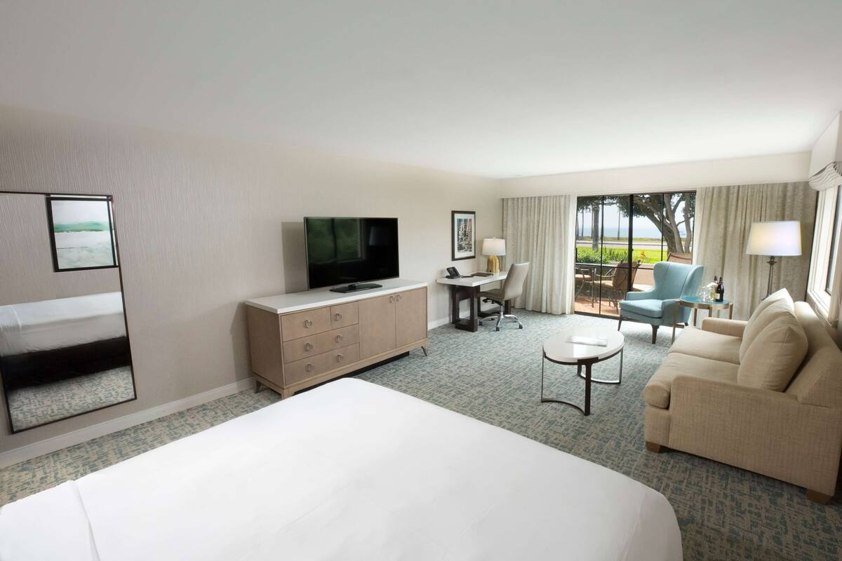 A hotel room with a large bed, a TV, a desk with a chair, a sofa, and a view of the outdoors.