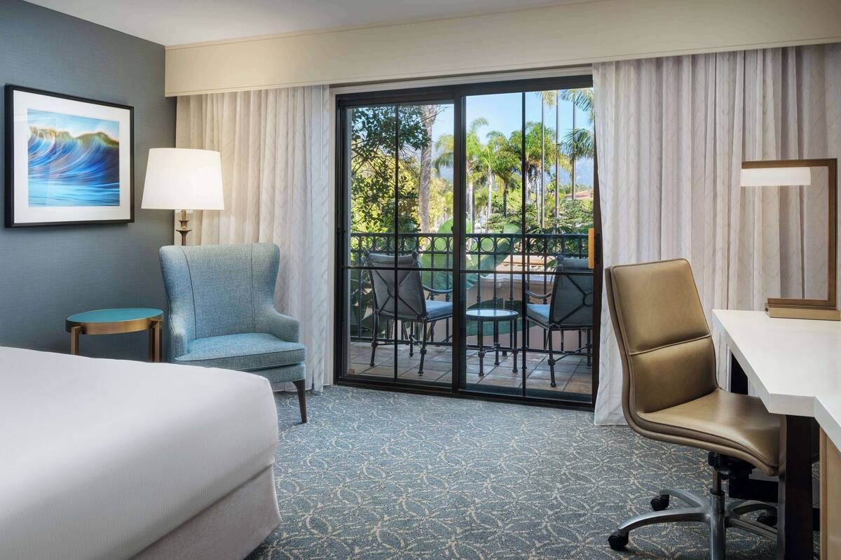 A hotel room with a bed, a desk, a chair, a small table, a lamp, and a view of a balcony with outdoor furniture.