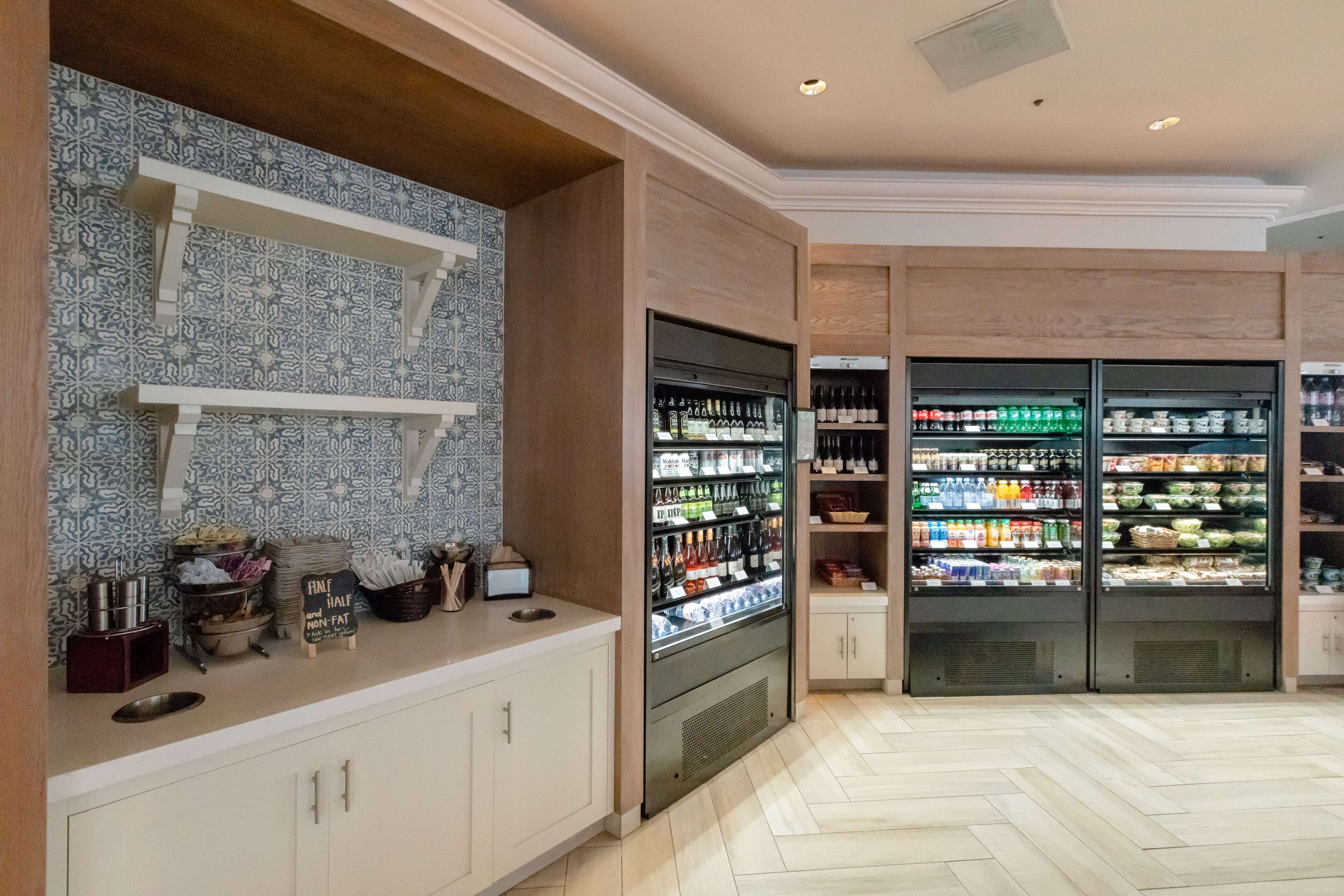 Une cuisine bien éclairée, avec un mur décoratif à motifs, un comptoir sur lequel se trouve une machine à café, et deux réfrigérateurs remplis de boissons et de produits alimentaires.