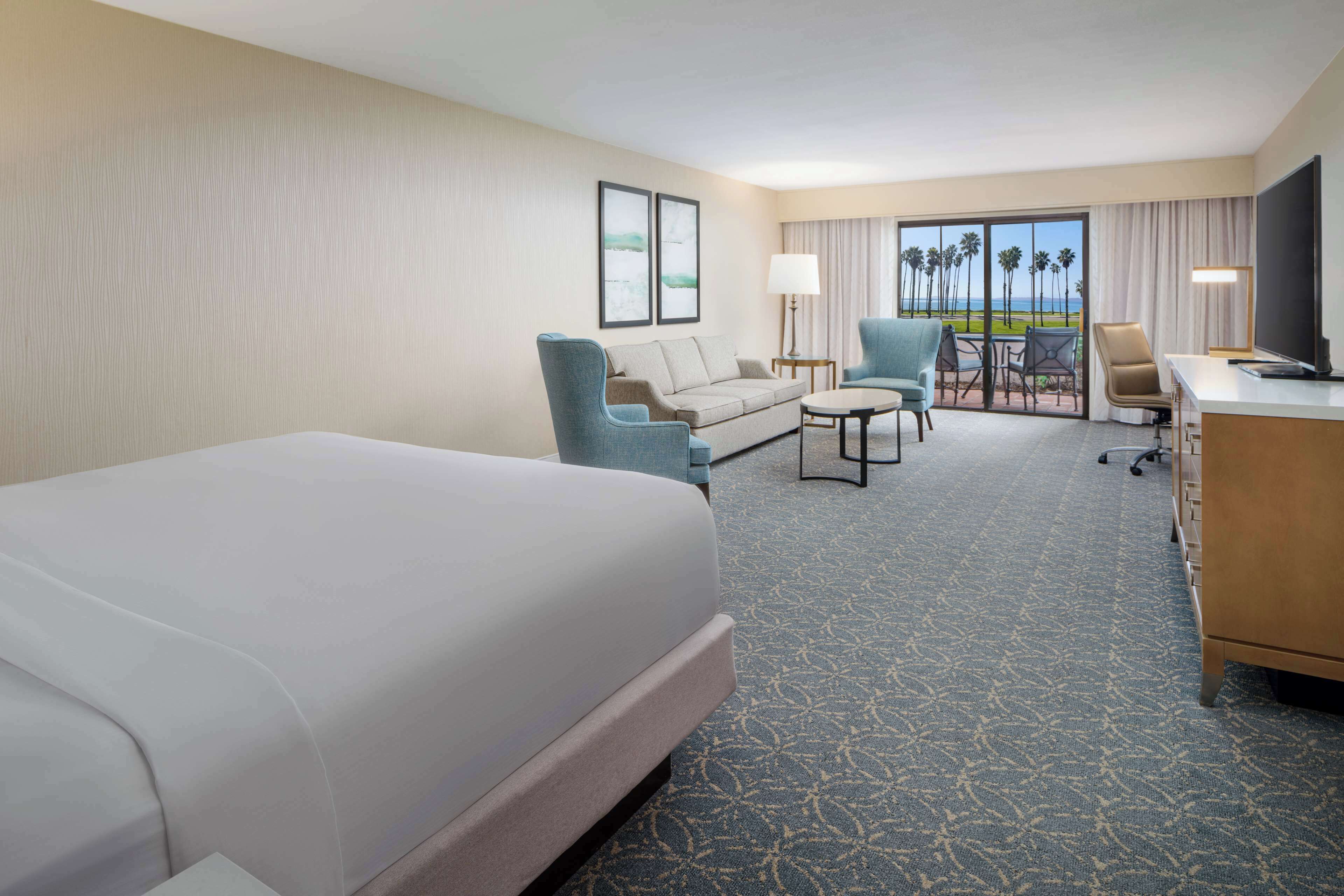 A hotel room with a bed, a sofa, a desk, and a chair. There is a view of palm trees and the ocean through the window.