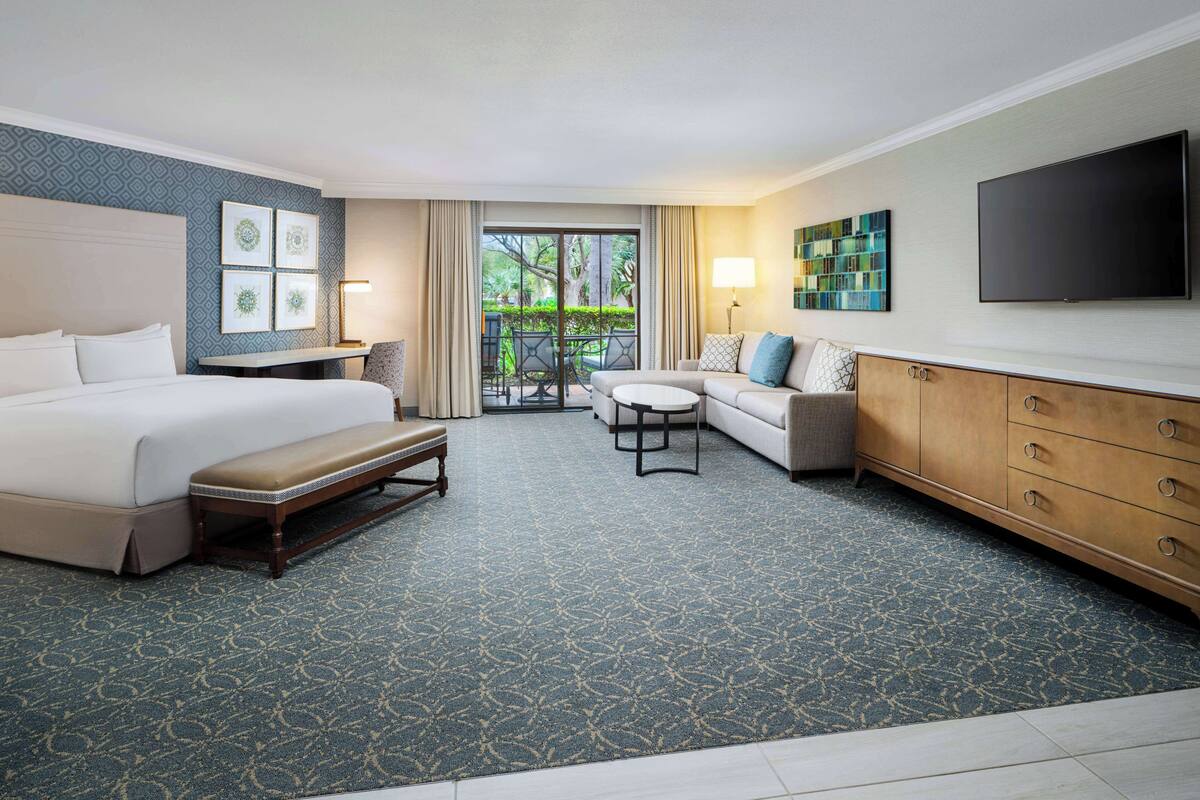 A hotel room with a bed, a sofa, a small table, a bench, a TV, and a balcony with a view of greenery.