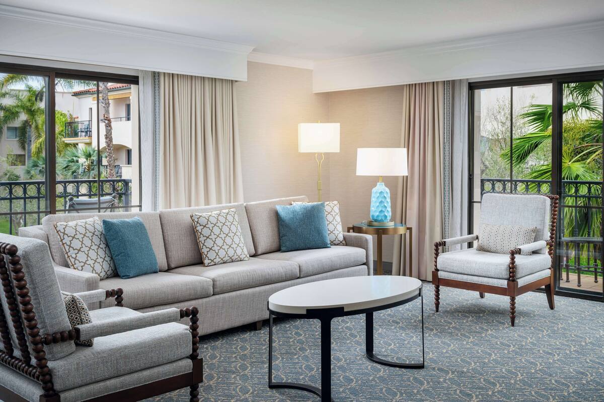 A hotel room with a beige sofa, a wooden armchair, a round coffee table, and a lamp.