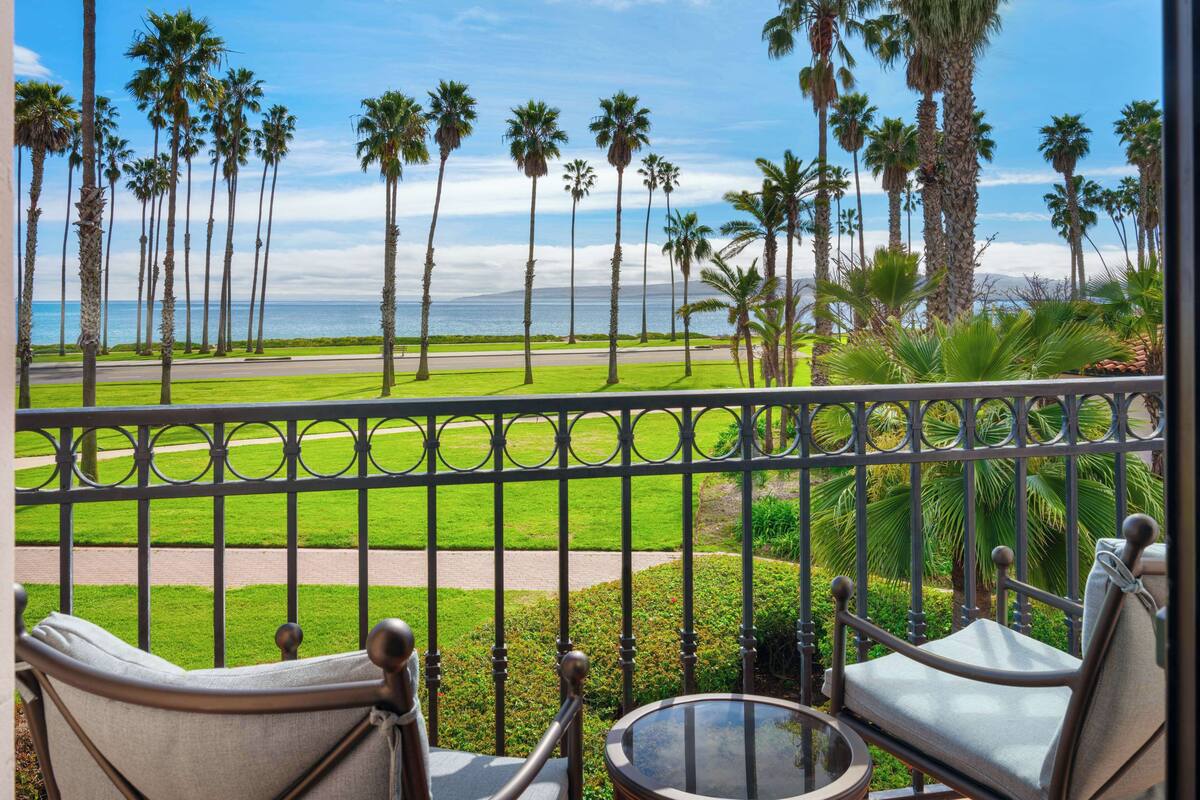 A balcony with two chairs and a small table, overlooking a well-maintained lawn and palm trees, with a view of the ocean.