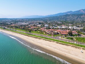 An aerial view of a coastal city with a beach, residential areas, and a mountainous backdrop.
