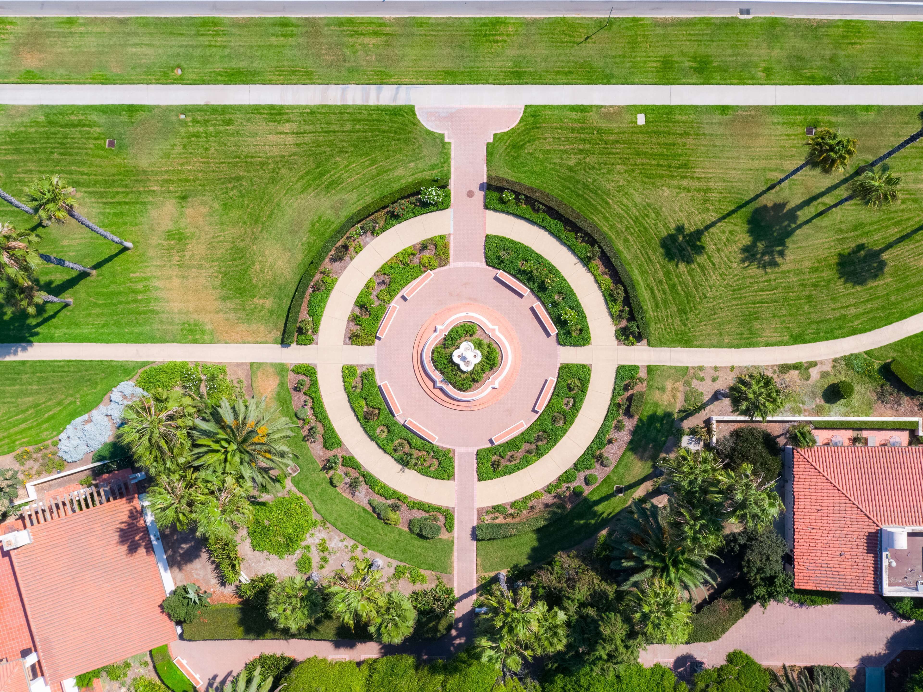 Vista aérea de um cruzamento ajardinado com um monumento circular central, cercado por vegetação e palmeiras.