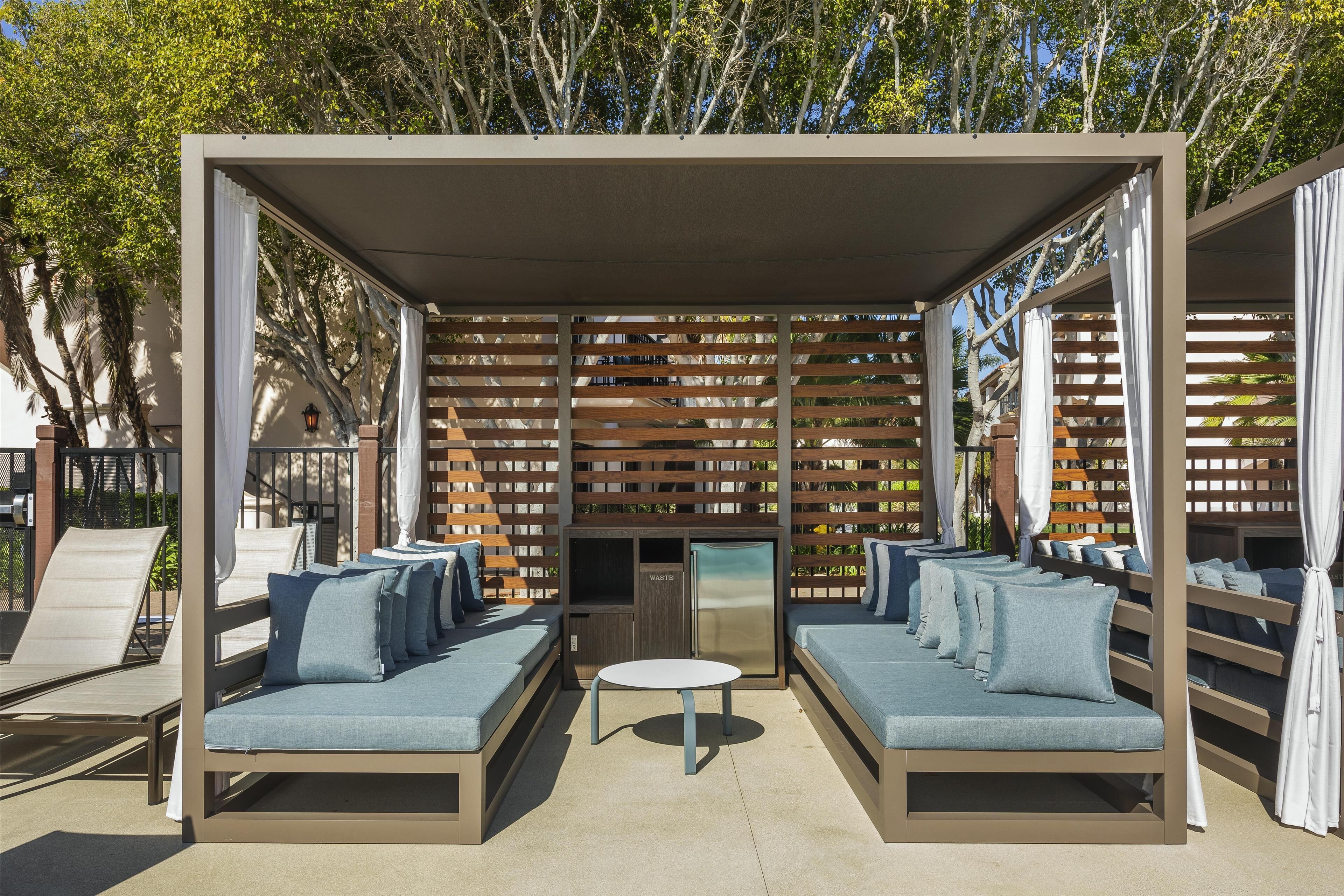 A covered outdoor seating area with cushioned benches and a central table, surrounded by wooden lattice panels.