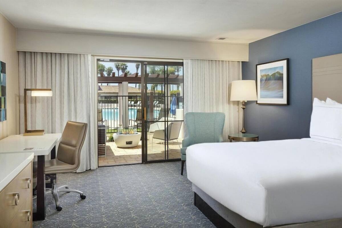 A hotel room with a bed, desk, chair, and a view of a pool through a glass door.