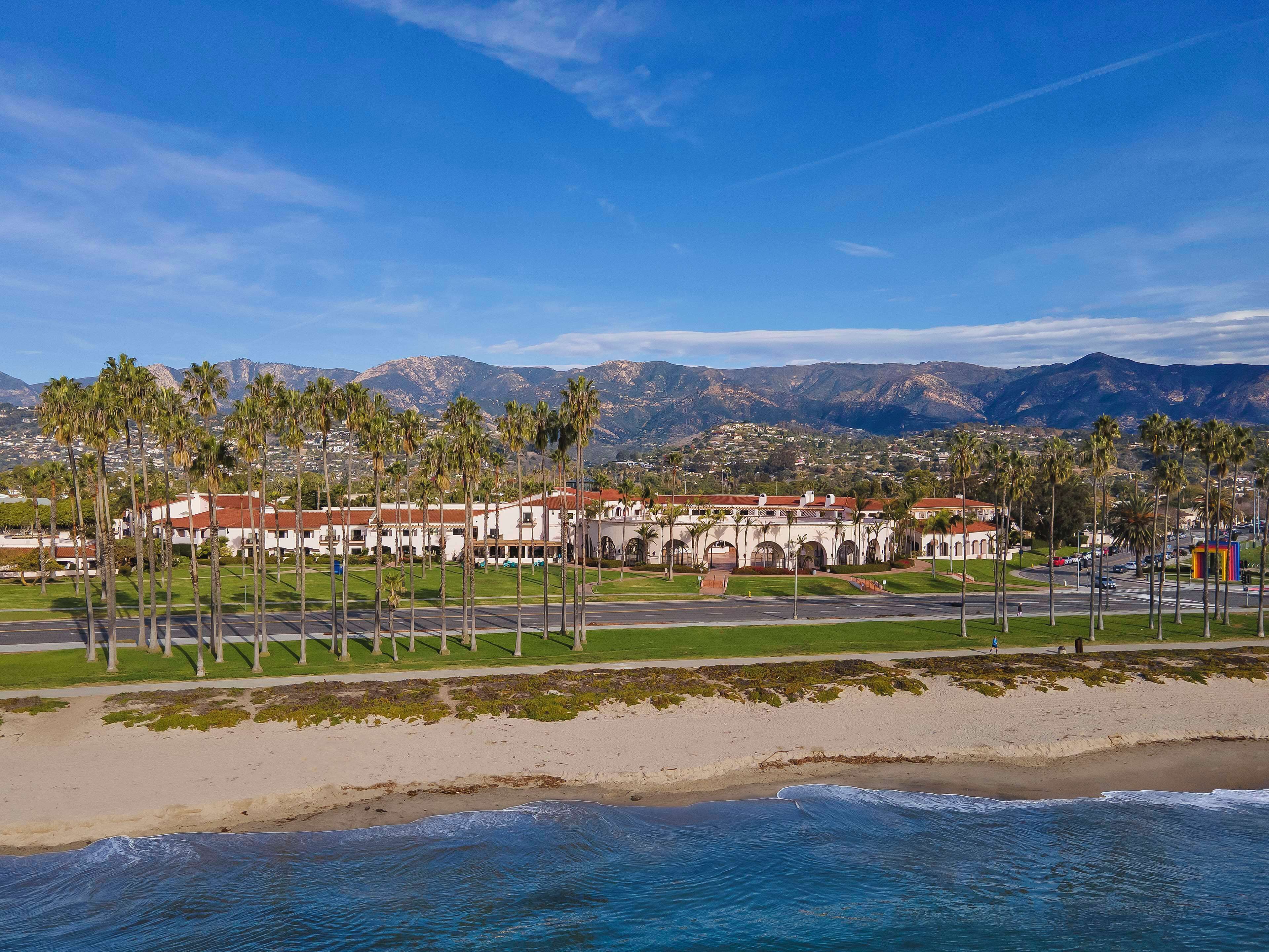 A coastal hotel with a beachfront view, palm trees, and a mountainous backdrop.