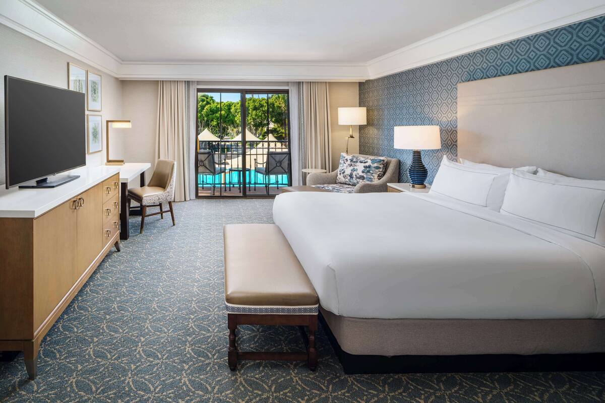 A hotel room with a large bed, a TV, a desk, a chair, a bench, and a view of a pool and greenery outside.