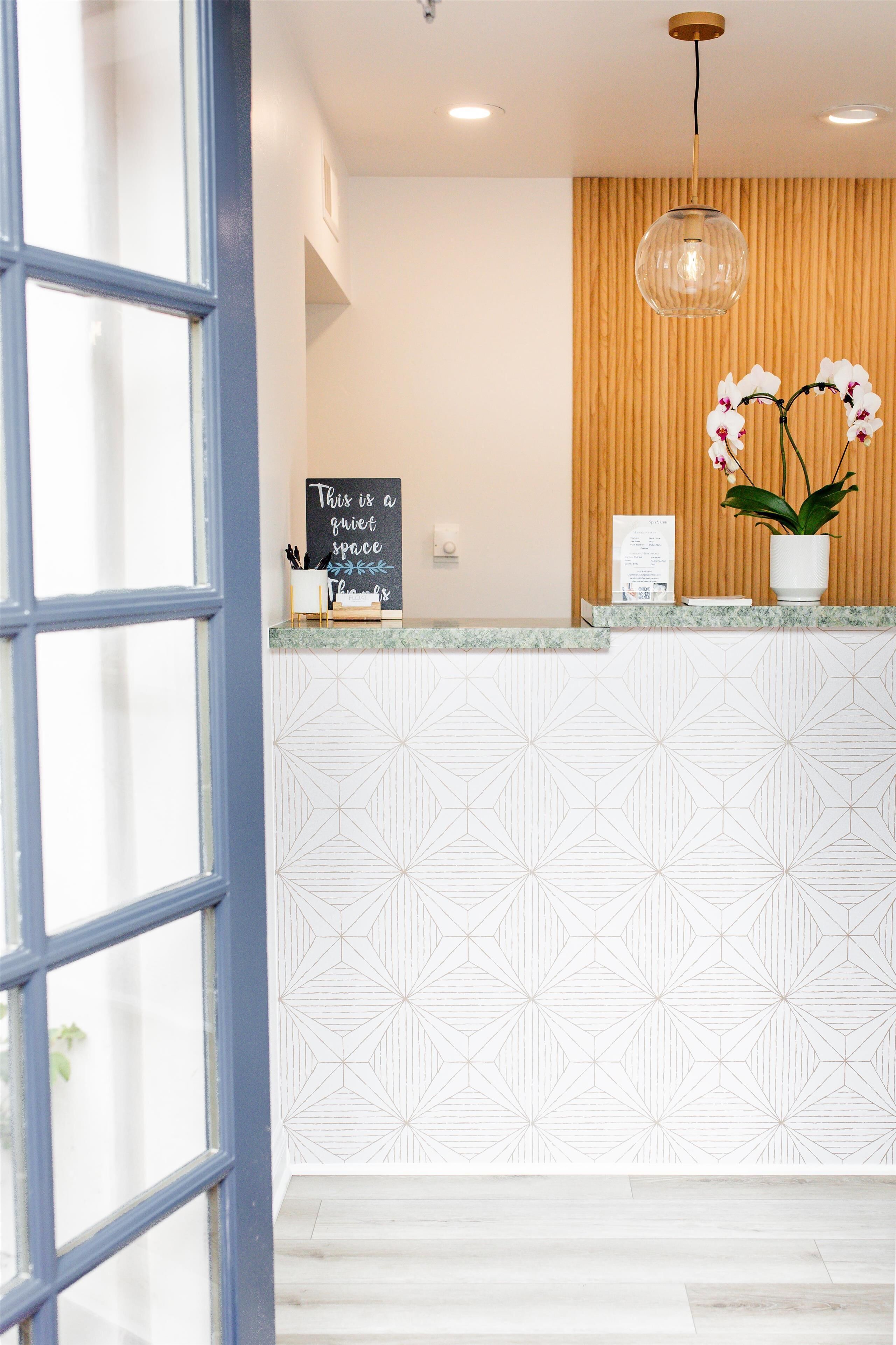 A reception area with a geometric patterned wall, a wooden panel wall, a hanging light fixture, a potted orchid, and a chalkboard sign.