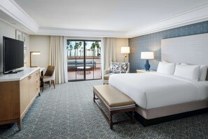 A hotel room with a large bed, a desk with a TV, a chair, a bench, and a view of palm trees through the glass door.