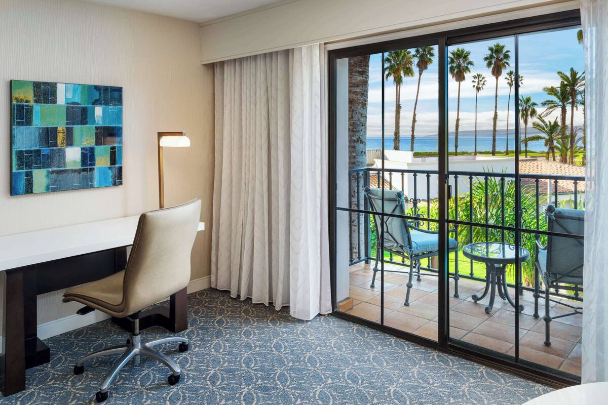 A hotel room with a desk, chair, and a view of a balcony with outdoor furniture and palm trees.