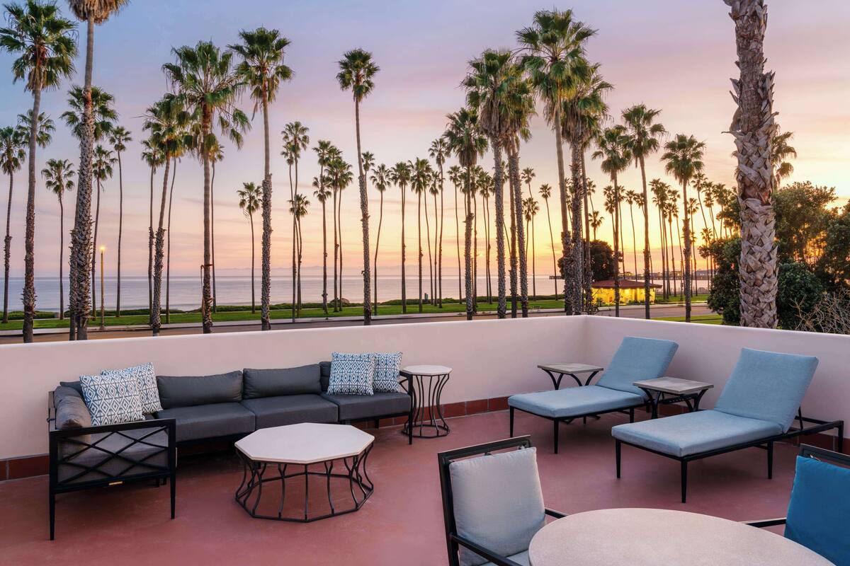 A terrace with a sofa, chairs, and tables, overlooking a beach with palm trees and a sunset.