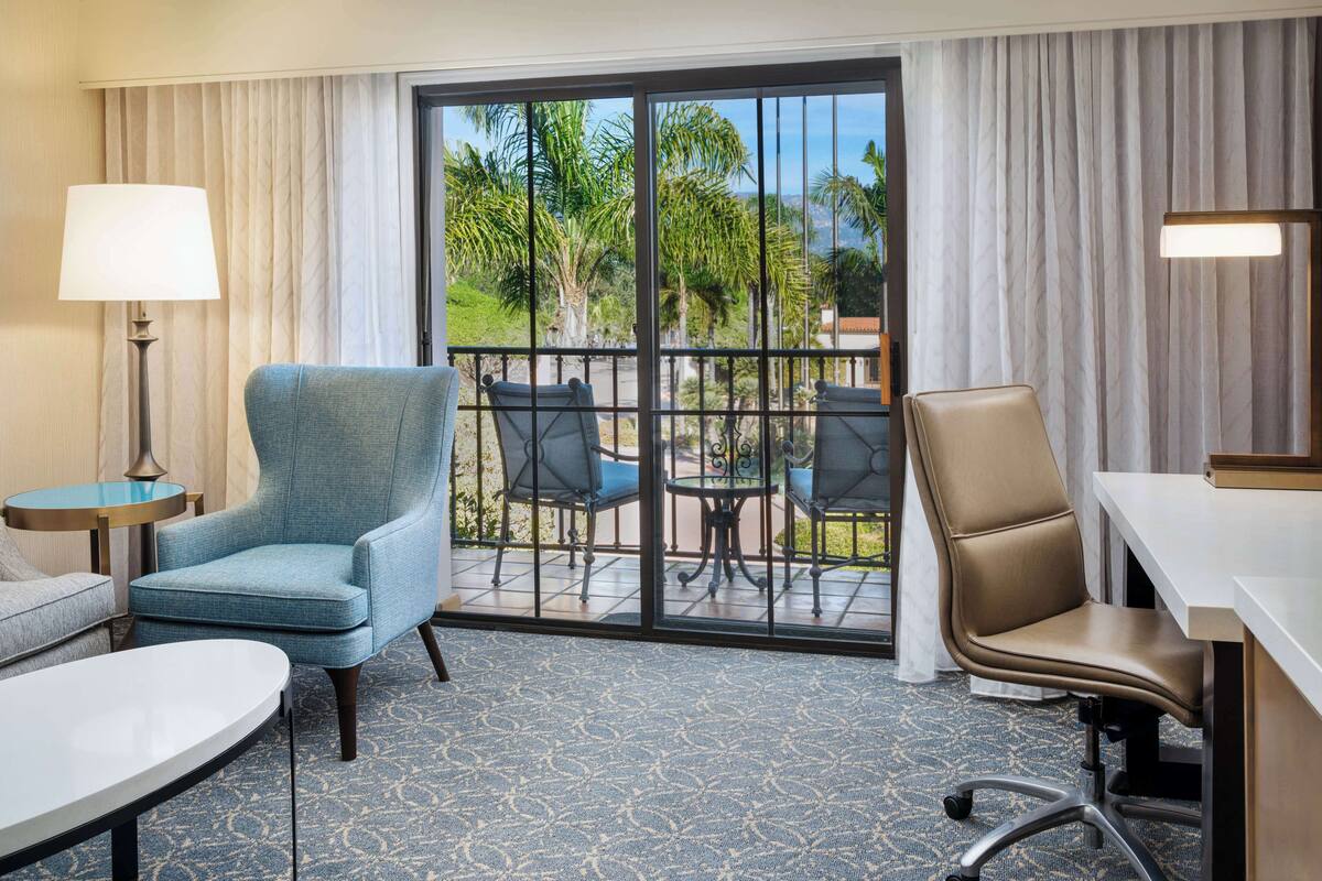 A hotel room with a blue armchair, a beige office chair, a small round table, a lamp, and a view of palm trees through a sliding glass door.