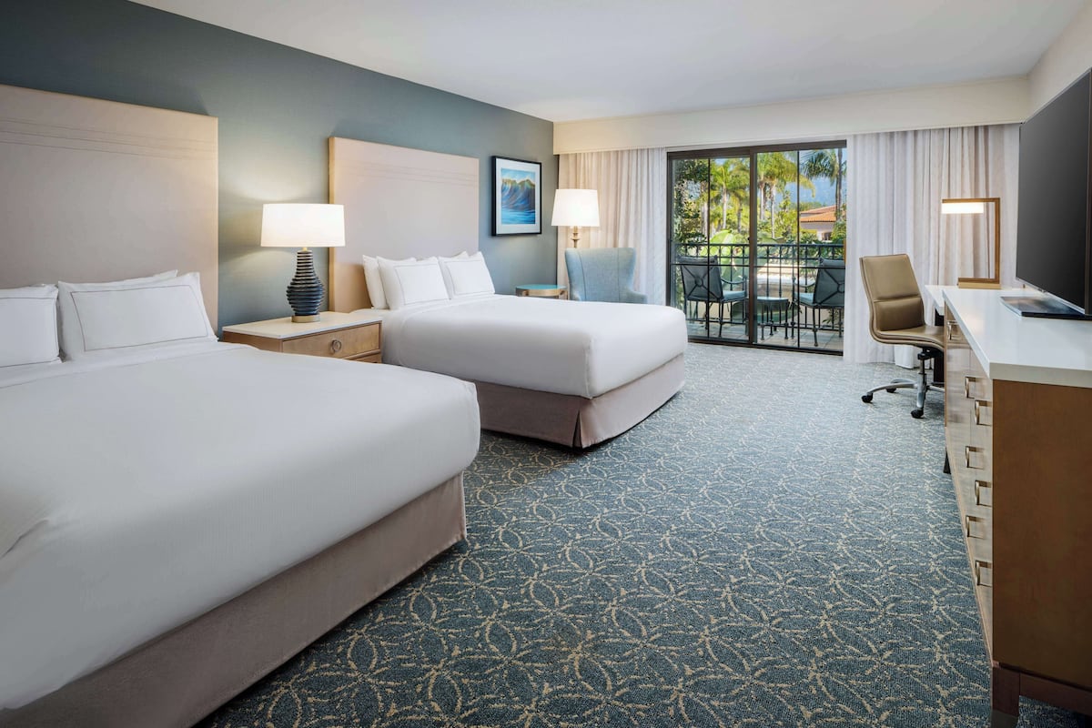 A hotel room with two beds, a desk, a chair, a laptop, and a balcony with a view of palm trees.